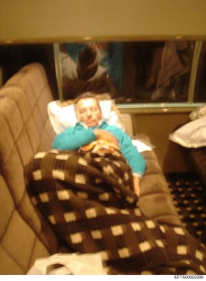 This is a candid photograph showing a man resting on a beige/tan upholstered couch wearing a turquoise/teal colored long-sleeved shirt. He appears to be lying back against white pillows with a brown and white patterned blanket covering his lower body
