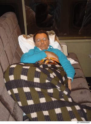 This is a candid photograph showing a middle-aged man resting on an aircraft seat, wearing a bright blue jacket and glasses. The image appears to be taken inside a private aircraft cabin, evidenced by the window visible in the background and the upho