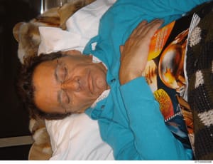 This is a photograph showing a person wearing glasses and a turquoise/blue cardigan over a white shirt, holding what appears to be a magazine or publication with orange and yellow imagery on its cover. The photo is taken indoors in low lighting condi