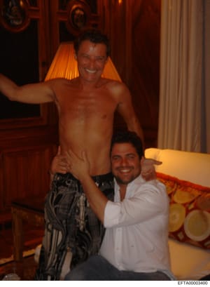 This is a candid photograph taken inside a luxury residence, showing two men posing together in what appears to be a living room or lounge area with ornate wooden doors, cream-colored furnishings, and warm amber lighting. The photograph is evidence d