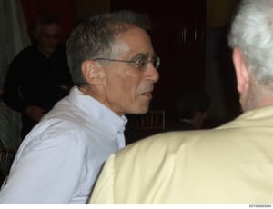 This is a photograph showing a middle-aged man wearing glasses and a light blue/white button-down shirt engaged in conversation with another person at what appears to be an indoor social gathering or event. The image quality and setting suggest it ma
