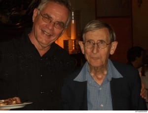 This is a photograph of two men at what appears to be an indoor social event or gathering. The image shows them posing together in a warm, convivial setting with warm lighting and dark wood paneling in the background. The photo's presence in the inve
