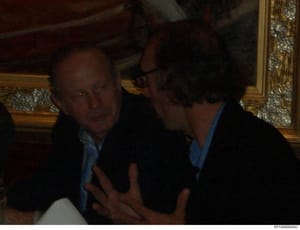 This photograph shows two men engaged in conversation in what appears to be an ornately decorated interior space. The setting features gold-trimmed frames and formal furnishings, suggesting an upscale residence. The photo quality and color characteri