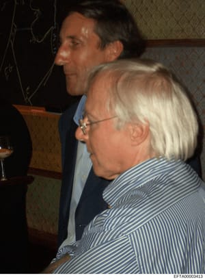 This is an evidence photograph from the Epstein investigation files showing two people in what appears to be an indoor social setting. The photo depicts a man in a dark blazer and light blue shirt standing behind a woman wearing a blue and white stri
