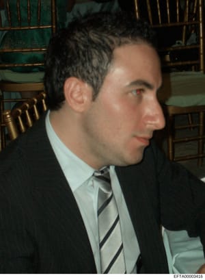 This is a photograph of an unidentified man in formal business attire (dark suit, light blue shirt, striped tie) photographed in profile at what appears to be an upscale interior setting. The photo shows the subject seated with decorative chairs with