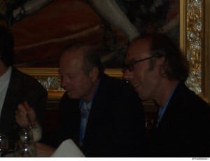 This photograph shows two men in dark suits and blue dress shirts engaged in conversation in an elegant interior space with ornate gold-framed mirrors and burgundy upholstered furniture visible in the background. The setting appears to be a formal so