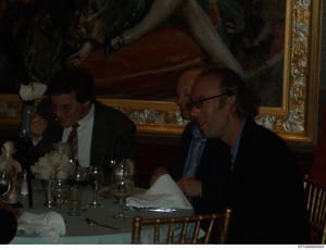 This is a photograph of what appears to be a formal dinner event in an ornately decorated room. Multiple people are seated at a dining table with place settings, glasses, and napkins. The setting suggests an upscale venue with decorative molding and 