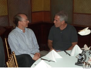 This photograph shows two men engaged in conversation at a formal dining table, likely during a business meeting or social gathering. The setting appears to be an upscale interior with decorative wallpaper paneling, and the table is set with place se