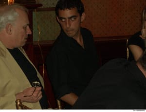 This is an evidence photograph showing two men in what appears to be an interior room with formal decor. One man wears a light-colored (cream/tan) jacket with dark shirt, the other wears a black shirt. The setting suggests a residence or formal venue