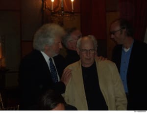 This photograph shows four men engaged in conversation at what appears to be an indoor social or professional gathering. The image quality is low and the lighting is dim, making definitive identification difficult. The setting suggests a formal or se
