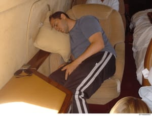 This is a photograph taken inside what appears to be a private aircraft cabin, showing a male subject sleeping in a beige/cream-colored upholstered chair. The image captures the interior of the aircraft with wooden trim and multiple seats visible. Th