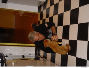 This is a personal photograph showing a man with gray hair wearing a black shirt and watch, holding a golden/tan-colored dog in an interior space with distinctive black and white checkered flooring and a red door. The photo appears to be taken indoor