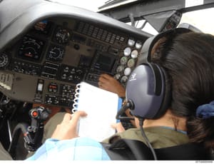 This photograph shows the interior cockpit of an aircraft with a pilot seated in the left seat, holding a spiral-bound notebook and wearing headphones. The image captures the aircraft's instrument panel, control yokes, and various flight instruments 