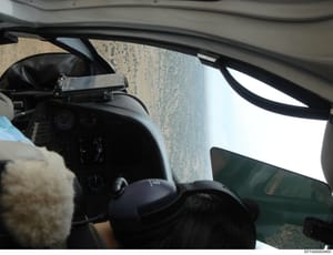 This photograph shows the interior of a vehicle taken from the passenger side, capturing the dashboard, steering wheel, windshield, and a view of what appears to be a beach or sandy coastal area. The image is rotated 90 degrees and shows minimal deta