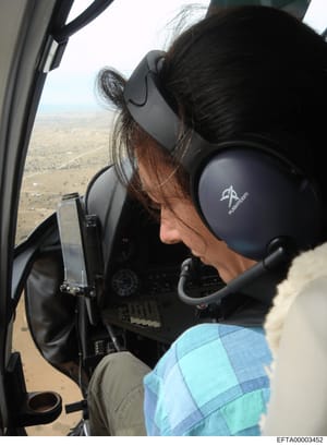 This photograph shows the interior of a helicopter during flight, with a passenger wearing aviation headset visible from behind. The image captures the cockpit environment, instrument panel, and aerial landscape below. The photograph appears to be do