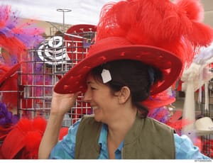 This is a photograph showing a person wearing a large red feathered hat in what appears to be a costume workshop or parade preparation facility. The image shows industrial scaffolding, mesh structures, and colorful decorative elements in purple and r