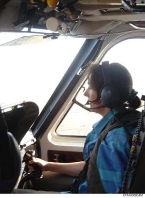 This is a photograph showing the interior of a helicopter cockpit with a pilot wearing a blue jacket and headset at the controls. The image appears to be taken from the passenger side, capturing the pilot in profile against a bright sky visible throu