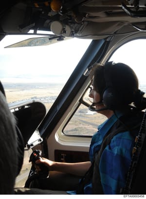 This photograph shows the interior of a small aircraft cockpit during flight, with a pilot or crew member visible wearing headphones and a blue flight suit. The image captures the instrument panel, controls, and a clear aerial view of rural landscape