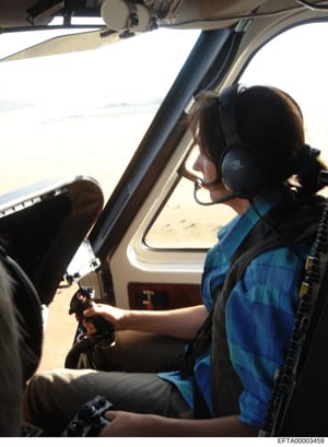 This is a photograph taken from inside a helicopter cockpit showing a person wearing a blue jacket and black headset seated in the co-pilot or passenger position. The image captures the interior of the aircraft with the windscreen visible and a lands