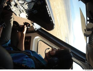 This photograph shows the interior view of an aircraft cockpit during flight, taken from the pilot's perspective. The image captures the instrument panel, controls, windshield, and desert landscape visible below, suggesting the aircraft was in flight
