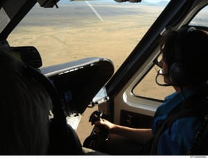 This is a photograph taken from inside an aircraft cockpit showing the view from the pilot's perspective over an arid, desert landscape. The image captures the interior of the aircraft with visible controls and instrumentation, along with a pilot or 