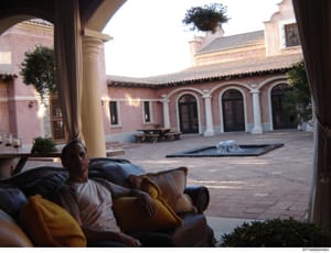 This photograph shows the interior courtyard of a substantial residential property with Spanish colonial-style architecture, featuring pink stucco walls, white columns, arched doorways, and decorative tilework. A male individual is visible in the for