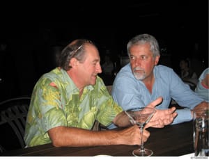 This is a photograph of two men engaged in conversation at what appears to be an evening social event or gathering. The men are seated at a table with drinks visible. The photo is undated and the identities of the individuals are not established from