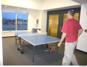 This photograph shows two men playing table tennis in what appears to be a high-rise building's recreation or game room. The room features floor-to-ceiling windows overlooking an urban cityscape at dusk, gold-colored elevators, and modern amenities. 