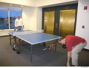 This photograph shows an interior recreation or break room in what appears to be a high-rise building, featuring two ping pong tables, two men engaged in play, large windows overlooking a cityscape at dusk, and two gold-colored elevators. The image i