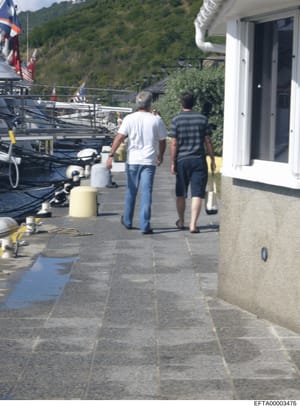 This photograph shows a waterfront dock area with a large boat moored on the left, a modern white building with glass doors on the right, and two men walking on a paved dock area. The setting appears to be a luxury waterfront property with forested h