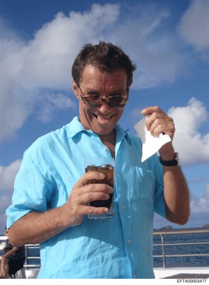 This is a casual photograph showing a middle-aged man on what appears to be a private boat or yacht, wearing a light blue shirt and sunglasses, holding a beverage and pointing at the camera. The photo was taken outdoors on a clear day with blue sky a
