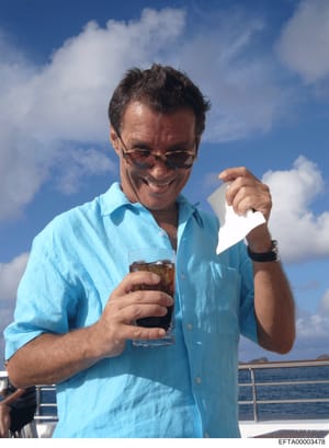 This is a color photograph of a middle-aged man aboard a yacht or boat, wearing a light blue button-up shirt and sunglasses, holding a glass with a dark beverage and a white napkin. The photograph appears to be personal/recreational in nature, taken 
