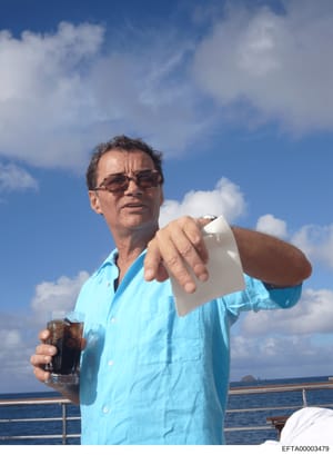 This is a photograph of an unidentified middle-aged man aboard what appears to be a yacht or large boat. The man is wearing a light blue casual shirt and sunglasses, holding a glass with a dark beverage and what appears to be a napkin or paper towel.
