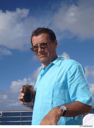 This is a color photograph showing a man on what appears to be a yacht or boat deck, photographed against a blue sky and ocean backdrop. The man is wearing a light blue short-sleeved shirt and sunglasses, holding a glass with a dark beverage. The pho