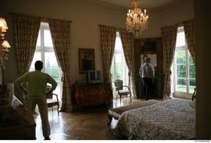 This photograph shows a large, elegantly appointed bedroom with multiple floor-to-ceiling windows draped in patterned curtains, a crystal chandelier, wooden furniture including a dresser and chairs, and hardwood flooring. Two individuals are visible 