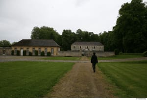 This photograph shows an expansive estate property featuring two substantial stone buildings on manicured grounds with a gravel drive and green lawn. The image captures the scale and architectural features of what appears to be a European country est