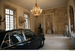 This photograph shows the interior of a large, elegantly decorated room with neoclassical architectural features, including ornate stone wall panels, classical sculptures, a gold chandelier, and framed windows. A dark vintage automobile is parked ins