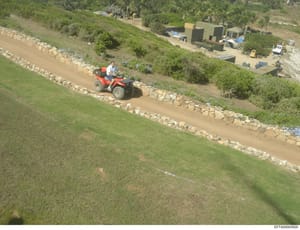 This is an aerial photograph showing a waterfront or coastal property under development or maintenance. The image captures a man on a red ATV working on a dirt path bordered by stone walls, with construction equipment, vegetation, and a modern struct