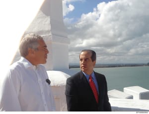 This photograph depicts two men in conversation on what appears to be a rooftop or elevated terrace overlooking a large body of water with coastal landscape visible in the background. One man wears a white shirt, the other wears a dark suit with a re