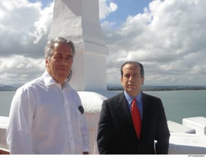 This is a photograph showing two men standing together at what appears to be a waterfront location with white architectural elements visible in the background. The image includes a scenic lake or water body with mountains in the distance under partly