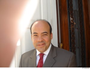 This is a photograph of a middle-aged man in professional business attire, taken indoors in what appears to be an upscale residential or office setting. The image shows the subject standing in front of ornate wooden architectural elements and white t