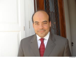 This is a portrait photograph showing a middle-aged male wearing a gray business suit with a white dress shirt and red plaid tie. The photo appears to be taken indoors in a formal setting with decorative wooden molding and curtains visible in the bac