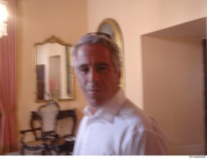 This is a photograph of a middle-aged man in a white shirt standing in an elegant residential interior space. The room features gold-framed mirrors, red curtains, and upholstered furniture, suggesting a high-end residence. The image appears to be par