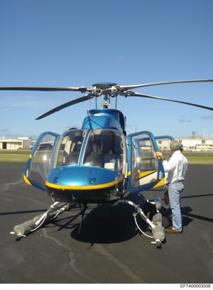 This is a photograph of a blue and yellow helicopter parked on an airport tarmac with its doors open and several people visible around it. The image appears to be documentation of helicopter operations or transportation assets, potentially relevant t