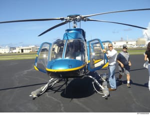This photograph shows a blue and yellow helicopter on an airport tarmac with several individuals standing beside and near the aircraft. The image appears to be documentation of helicopter operations or a charter flight, with clear visibility of the a