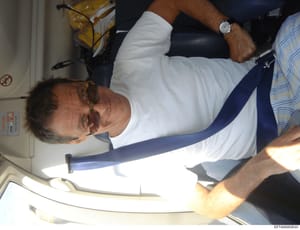 This is a photograph taken inside an aircraft, showing a man wearing sunglasses, a white shirt, and a dark blue tie seated in what appears to be a private plane cabin. The image is rotated and shows the interior environment of the aircraft with visib