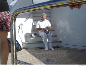 This is a photograph taken inside what appears to be a yacht cabin or luxury vessel interior. The image shows a gray-haired man in casual clothing (white t-shirt, light pants, white socks) sitting on wooden stairs inside the cabin. The photo captures