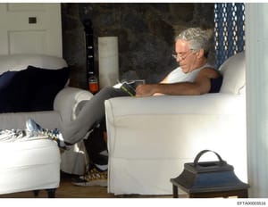 This photograph shows a man with gray hair and glasses sitting on a white upholstered chair/sofa in what appears to be a residential interior space. He is holding what looks like reading material. The room features dark marble or stone walls, blue pa