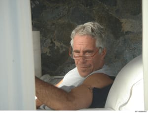 This is a color photograph showing a man wearing glasses and a light gray shirt, photographed indoors against a dark marble or granite background. The image appears to be an evidence photo or custody photograph from the investigation. The Bates numbe