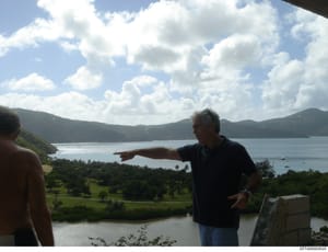This photograph shows an unidentified man in a dark shirt gesturing across a scenic tropical landscape featuring a large bay or body of water surrounded by forested hills and mountains. The image appears to be taken from an elevated vantage point on 